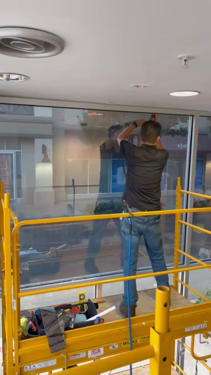 Installer on a yellow lift platform working film onto a tall interior window