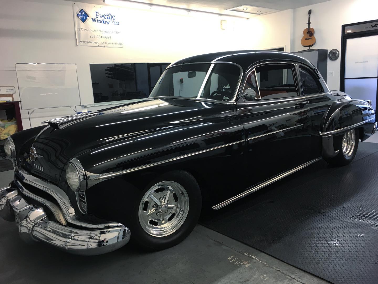 Black 1950s Oldsmobile coupe inside the Pacific Window Tint shop under the shop banner