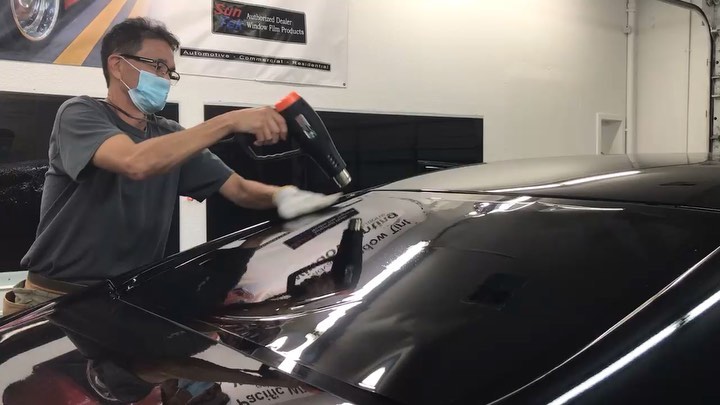 Installer using a heat gun and squeegee to lay paint protection film across the hood of a dark car